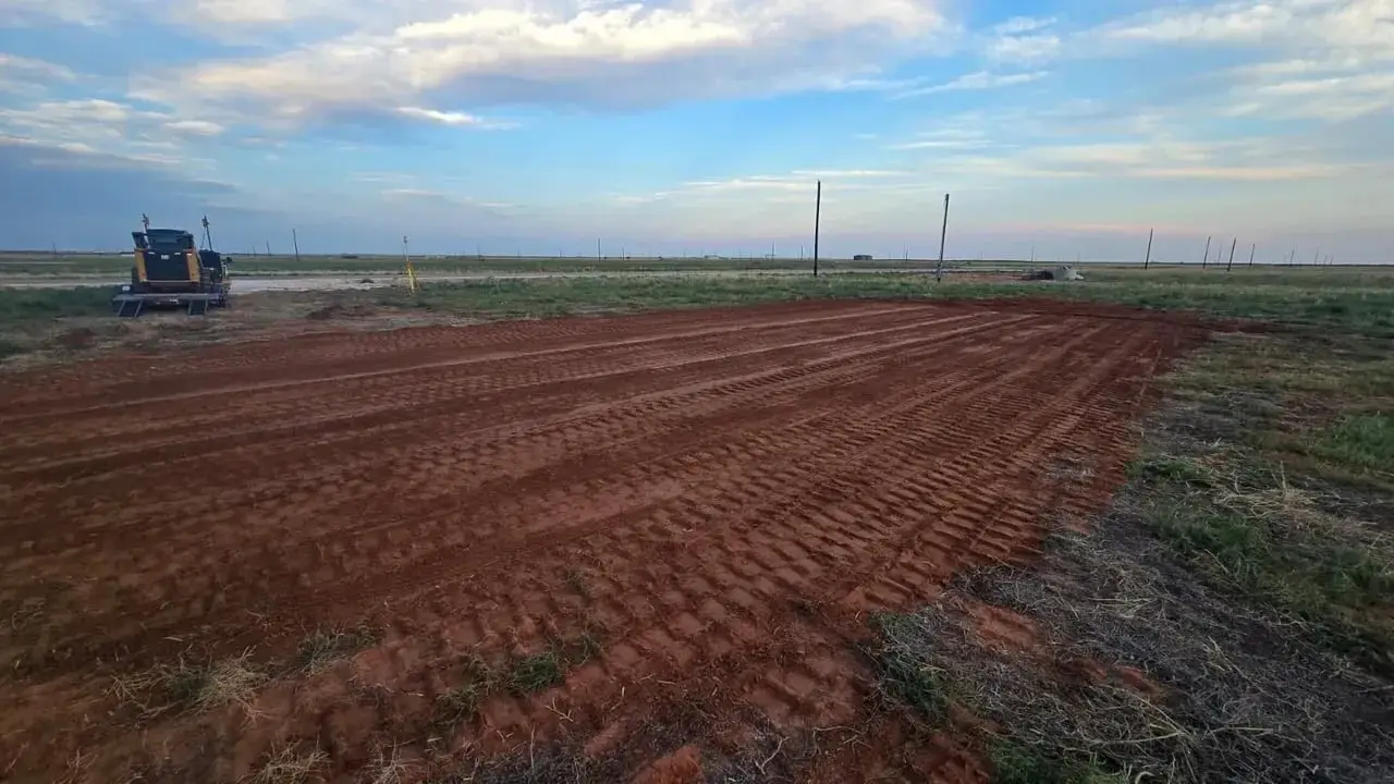 A cleared and rough-graded red dirt pad in an open field at dusk