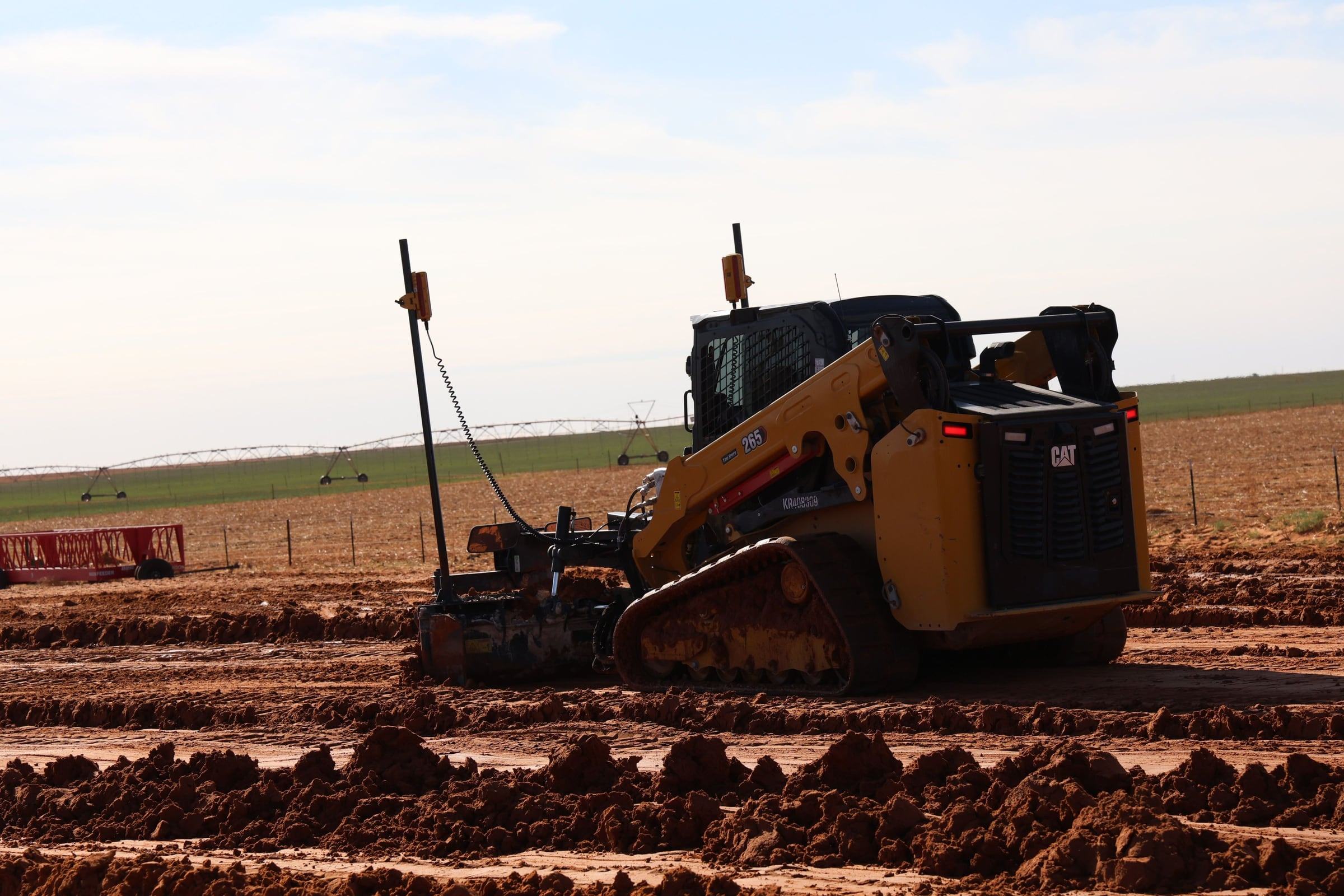 Track loader pulling a laser-guided grade box across red dirt on a West Texas job site at low sun