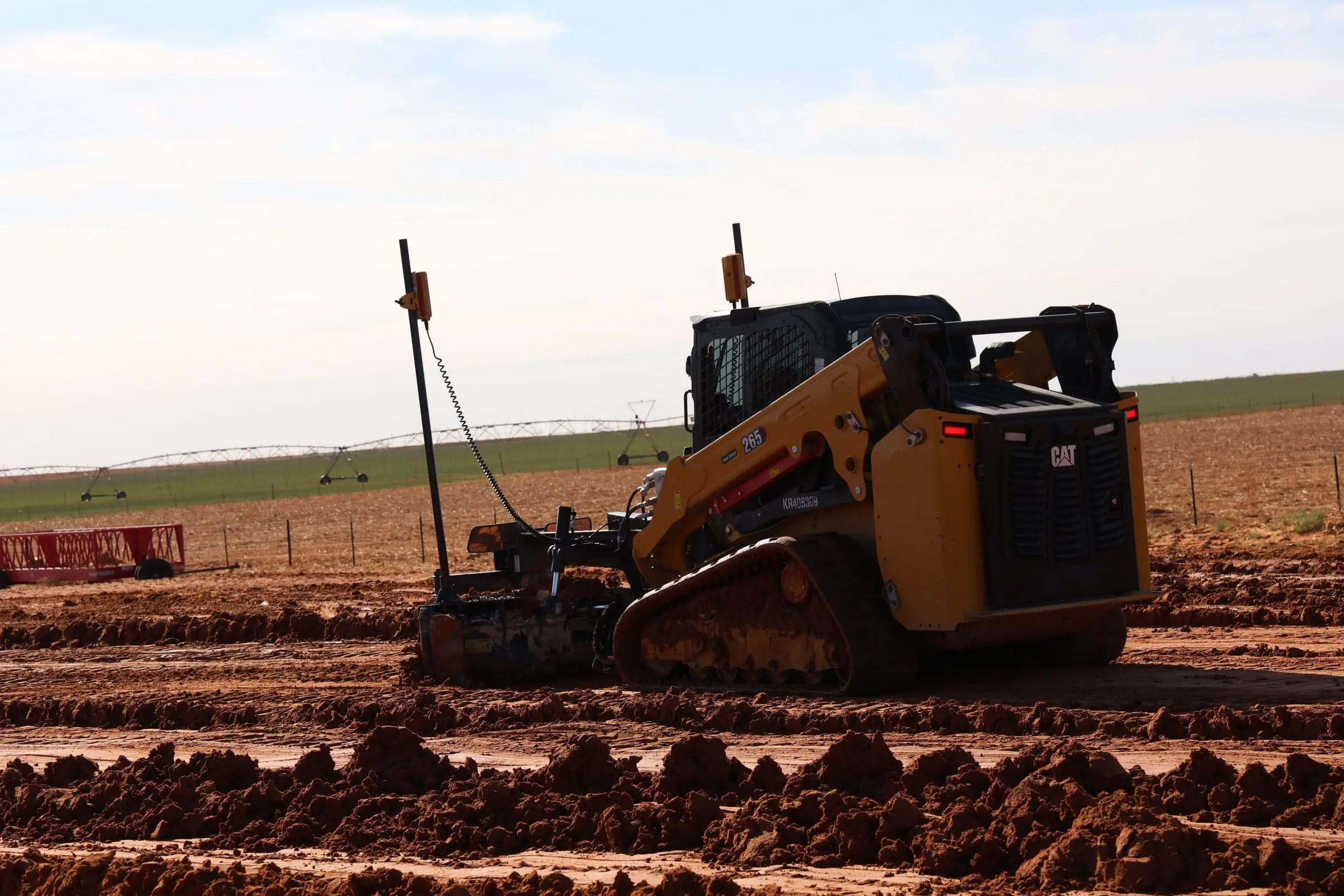 Track loader pulling a laser-guided grade box across red dirt at low sun