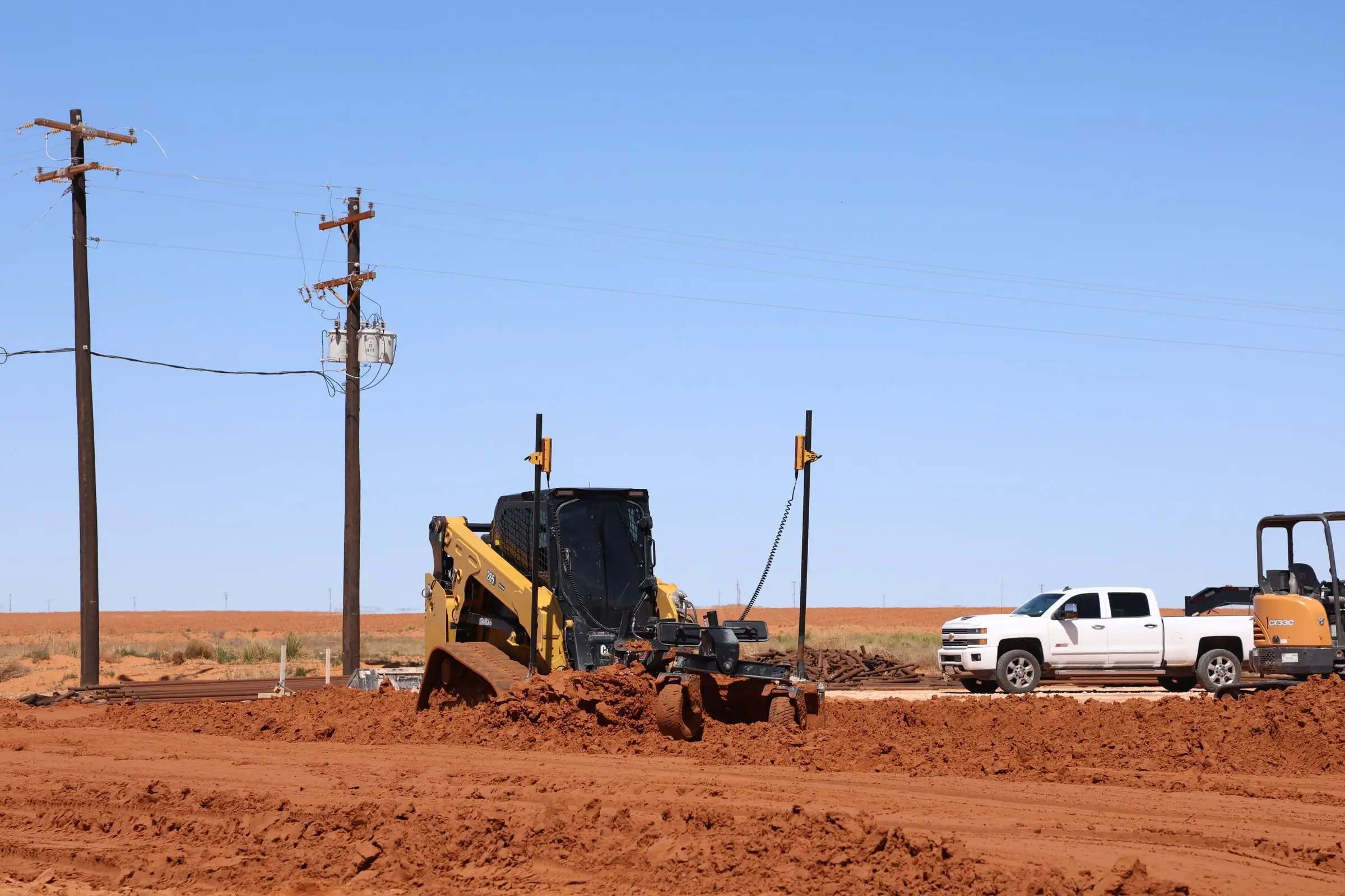 Skid steer grading a red dirt pad beside a pickup and a mini excavator