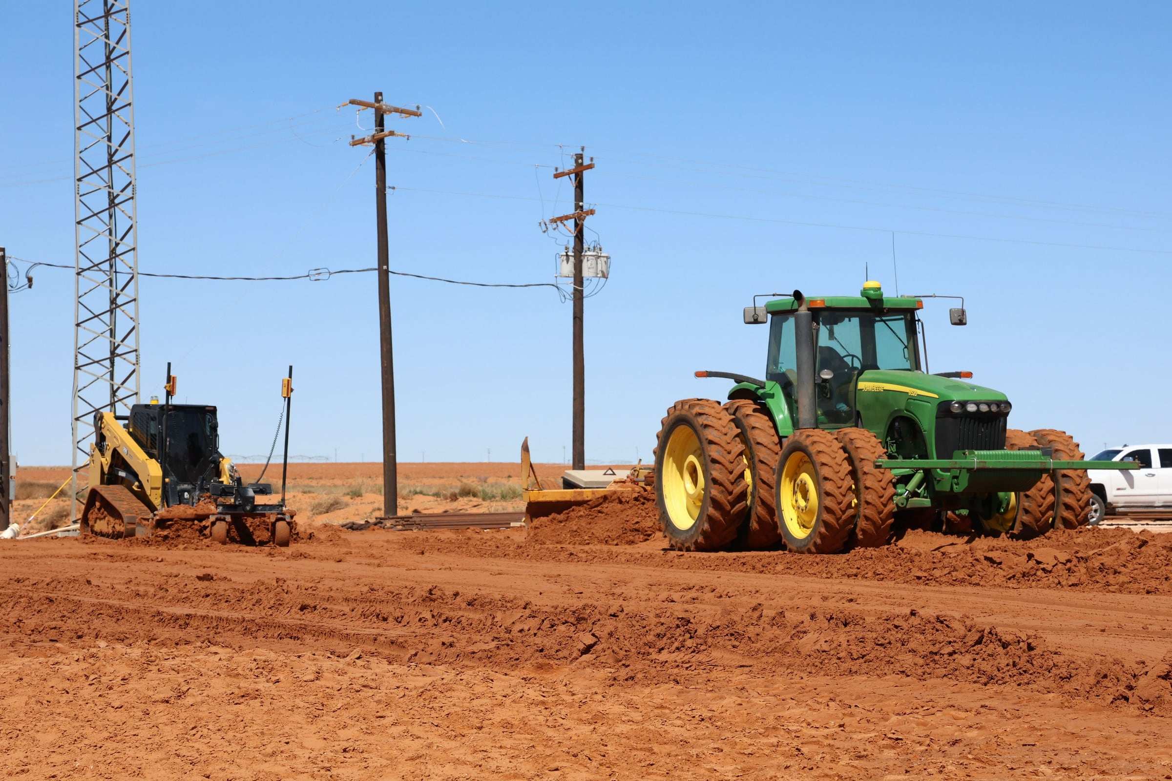 Farm tractor pulling a scraper alongside a compact track loader on a graded red dirt site